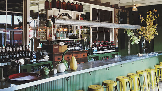 interior of a rustic bar with yellow metal barstools, a metal bar top, and cider bottles lined up along the wall behind the bar