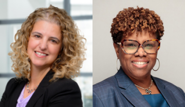 Two women in business attire smiling at the camera; one has curly blonde hair and a black blazer, the other has short curly hair, glasses, and a blue blazer, standing against neutral backgrounds.