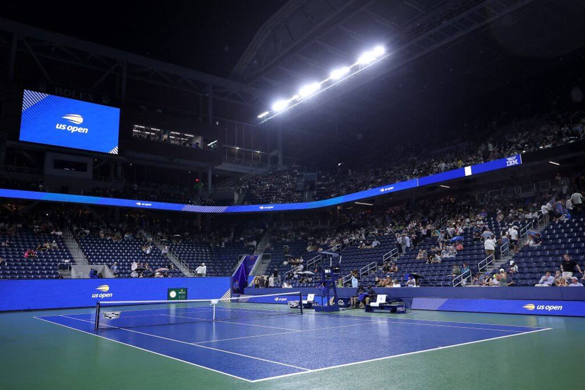 An open tennis court with rain on it and floodlights.