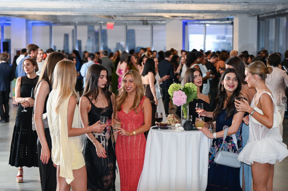 A crowded benefit reception shows guests gathered around cocktail tables with drinks and flowers in a large event space.