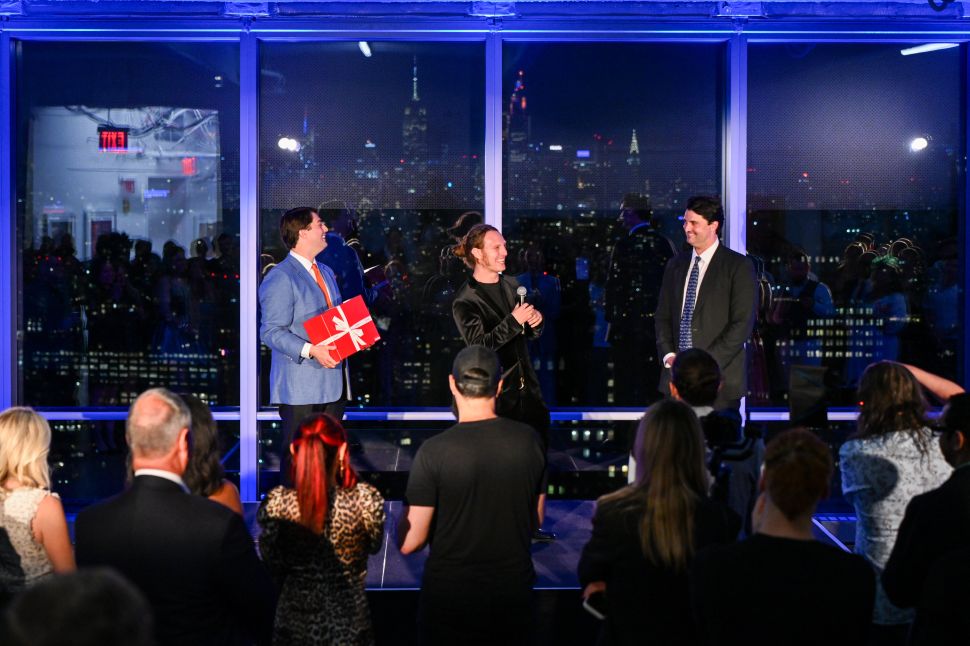 A speaker stands between two men at an evening benefit event, addressing a seated audience in front of floor-to-ceiling windows overlooking the New York skyline.