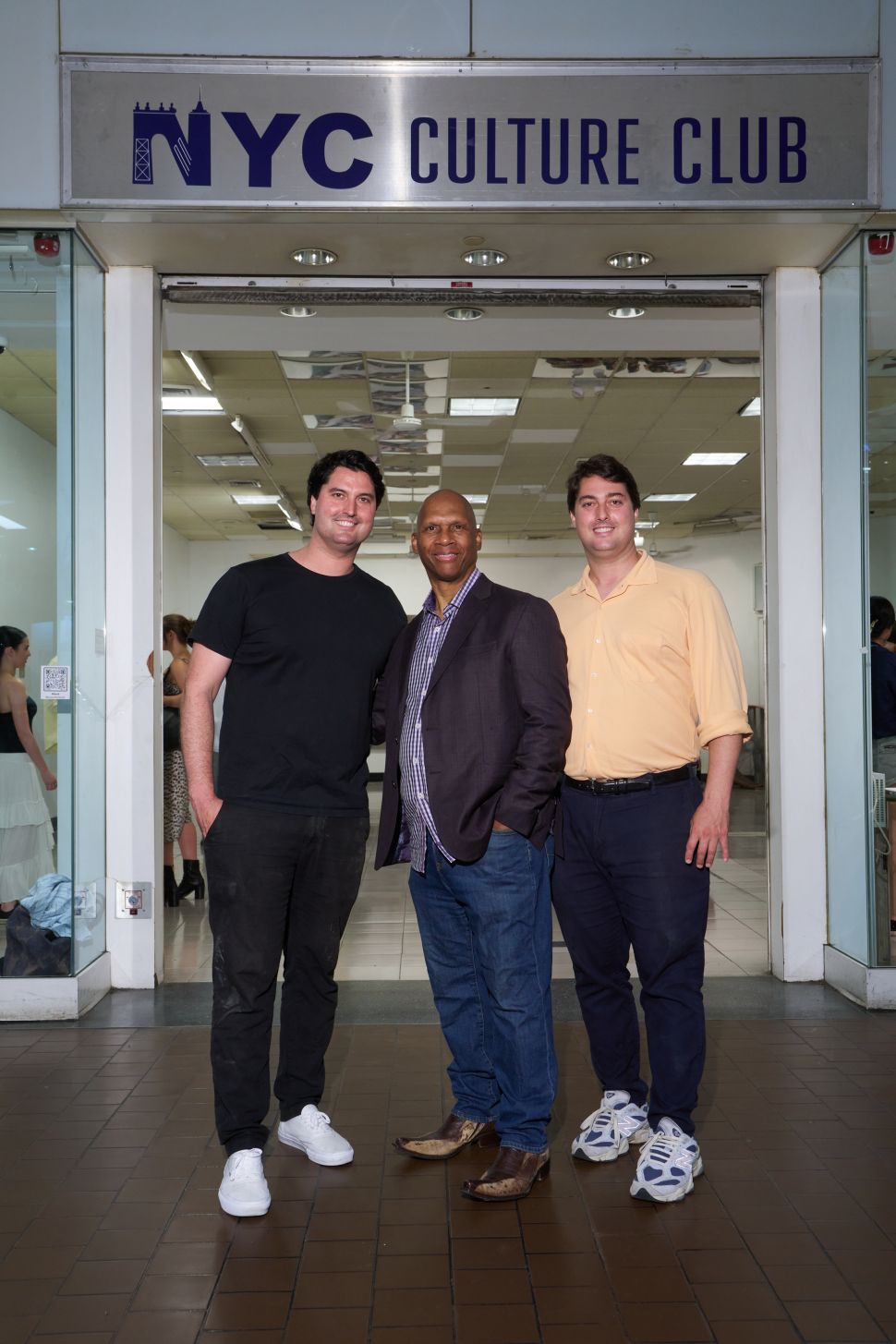 Three people pose beneath the NYC Culture Club sign at the entrance to a brightly lit gallery space inside a transit terminal.