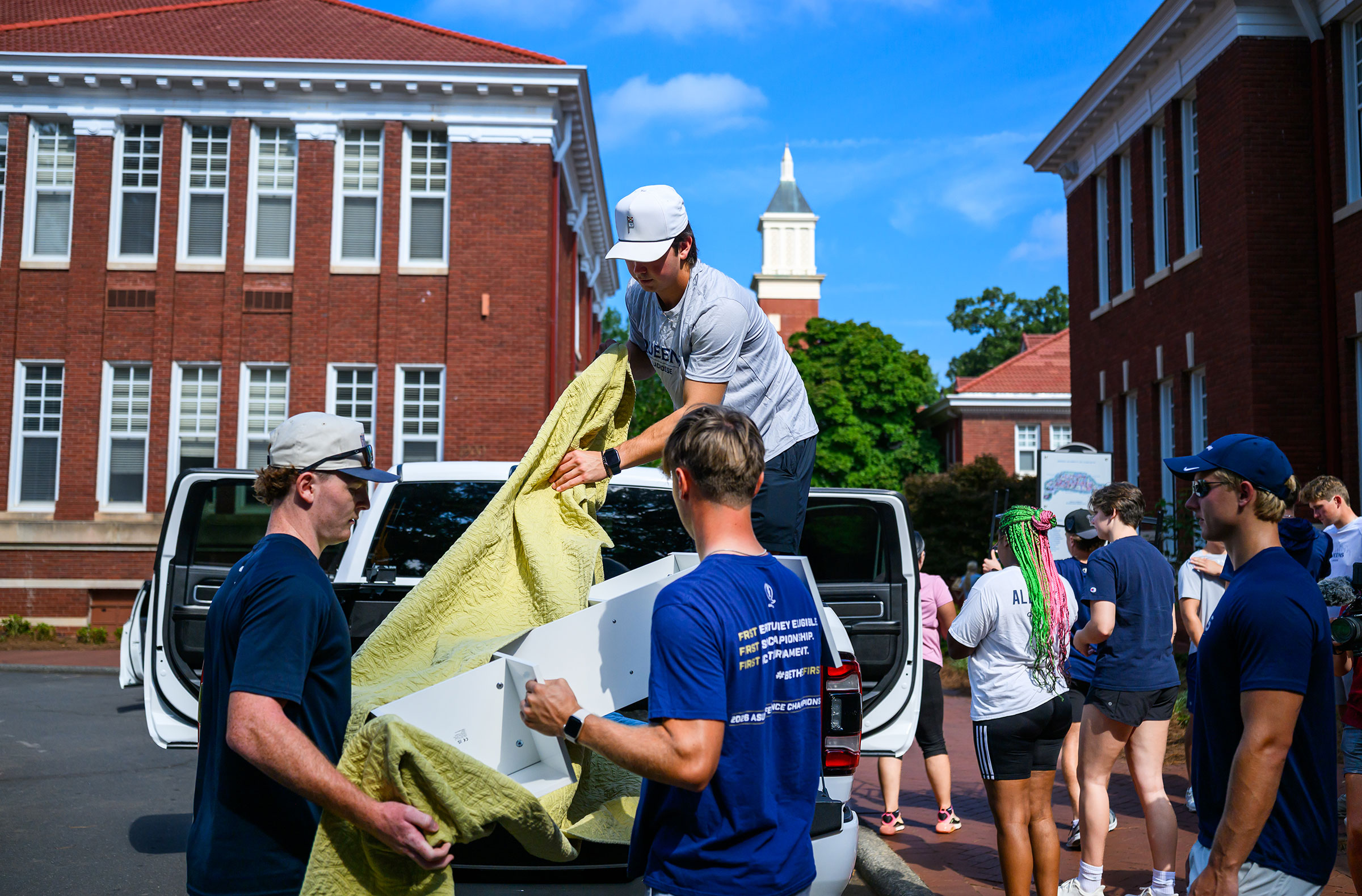 Men and women wearing blue Queens teeshirts unload furniture and bags out of a car in front of red brick buildings.