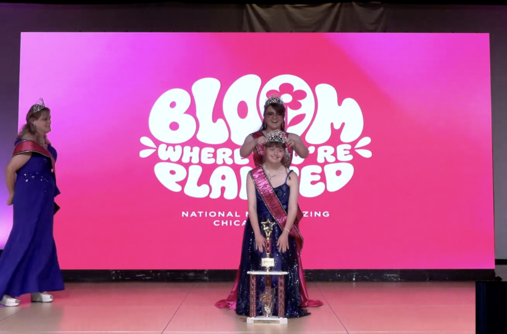 girl in a blue dress with a sash and trophy, with another woman putting a crown on her head.
