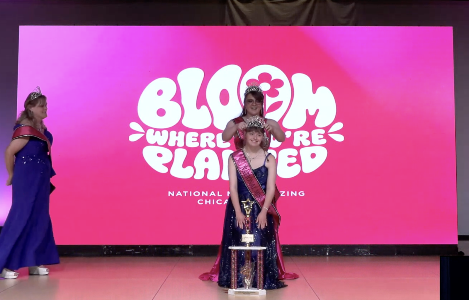 girl in a blue dress with a sash and trophy, with another woman putting a crown on her head.