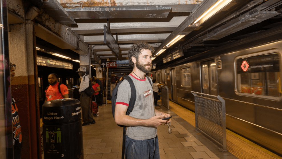 A young white man with curly brown hair and beard, with a backpack, stands on a subway platform