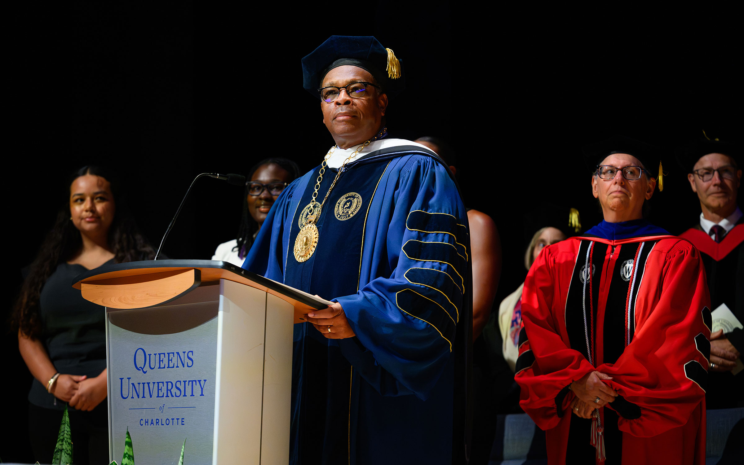 Man with glasses in blue regalia and cap stands at a podium marked Queens University of Charlotte.