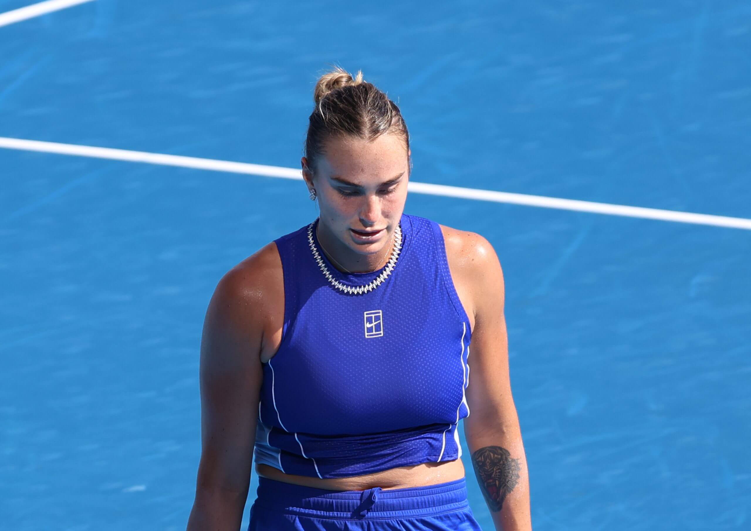 Aryna Sabalenka looks on with a blue tennis court behind her.