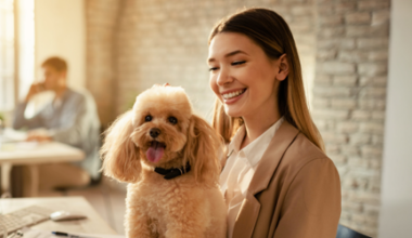 Smiling woman at a desk in an office pets a small, fluffy dog sitting on her lap. A computer screen and another person working are visible in the background.
