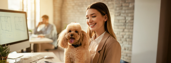 Smiling woman at a desk in an office pets a small, fluffy dog sitting on her lap. A computer screen and another person working are visible in the background.
