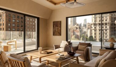 A neutral living room in a penthouse with a balcony running around it with a glass balustrade with two matching wood coffee tables in the center of mirroring armchairs and a sofa