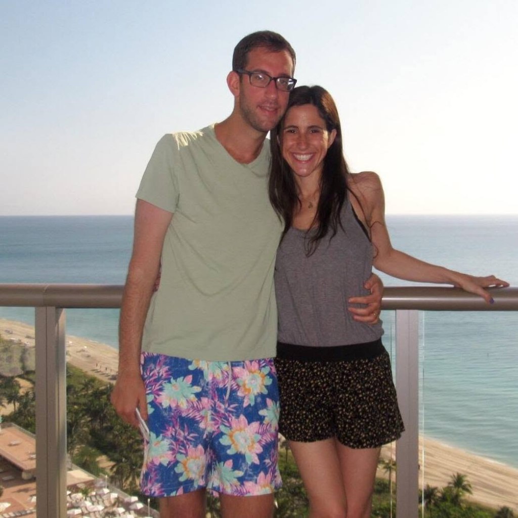 Allison Rahmani and her husband, Matthew, posing together on a balcony overlooking the ocean.