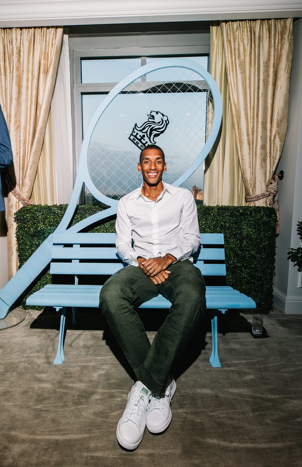 Smiling man sitting on a blue bench with a large tennis racket backdrop and indoor decor.