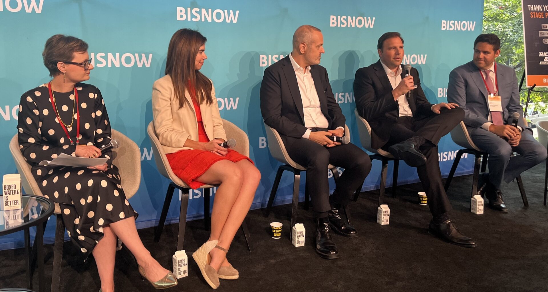 Five people seated on a stage speak into microphones at a Bisnow event. A blue backdrop with the Bisnow logo is behind them.