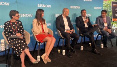 Five people seated on a stage speak into microphones at a Bisnow event. A blue backdrop with the Bisnow logo is behind them.