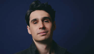 A man with dark hair and a slight smile wears a dark jacket, standing against a solid dark blue background.