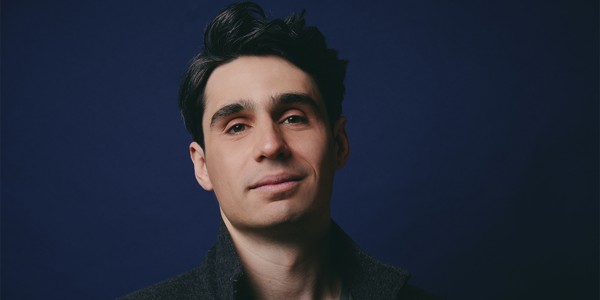 A man with dark hair and a slight smile wears a dark jacket, standing against a solid dark blue background.