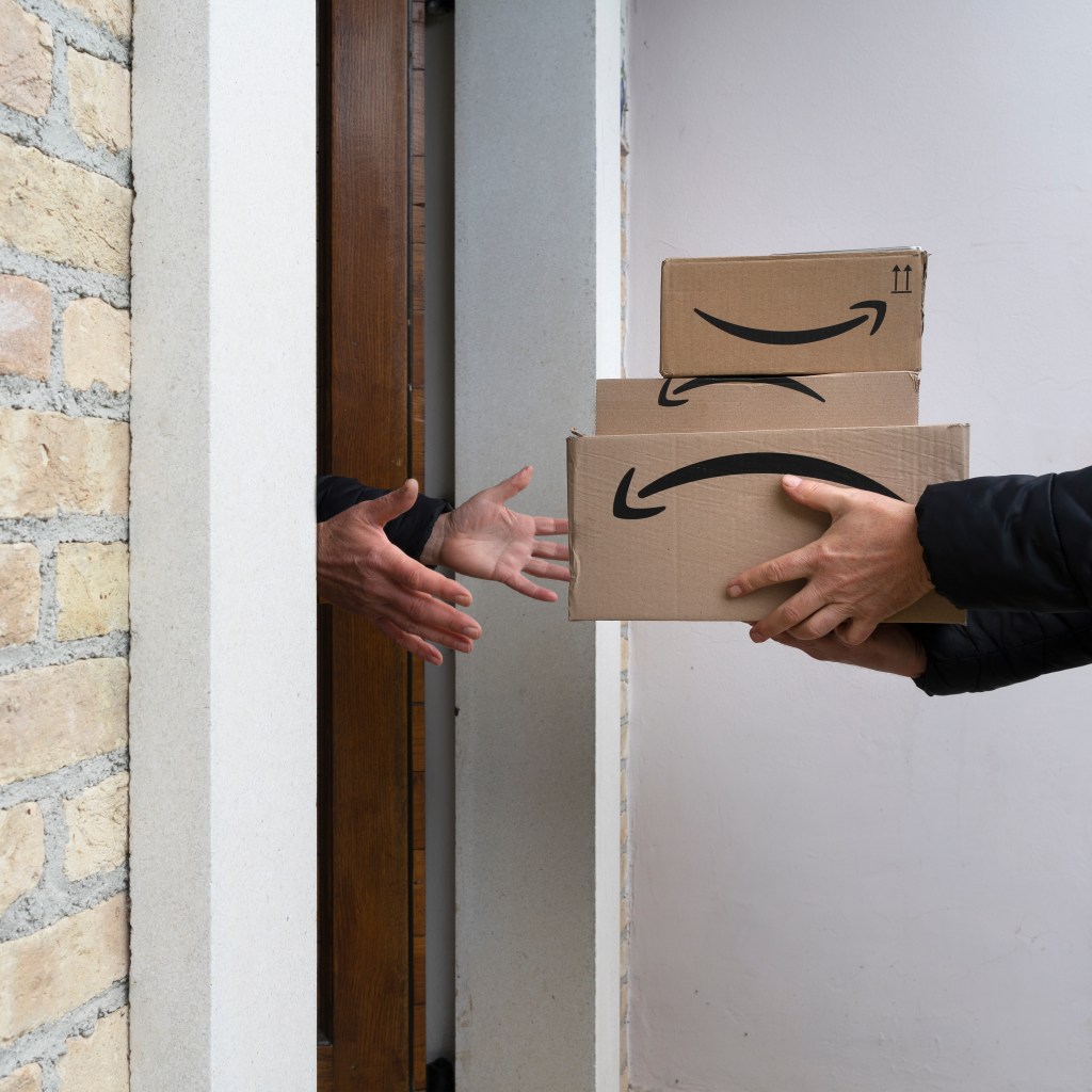 A person handing three Amazon boxes to another person through a doorway.