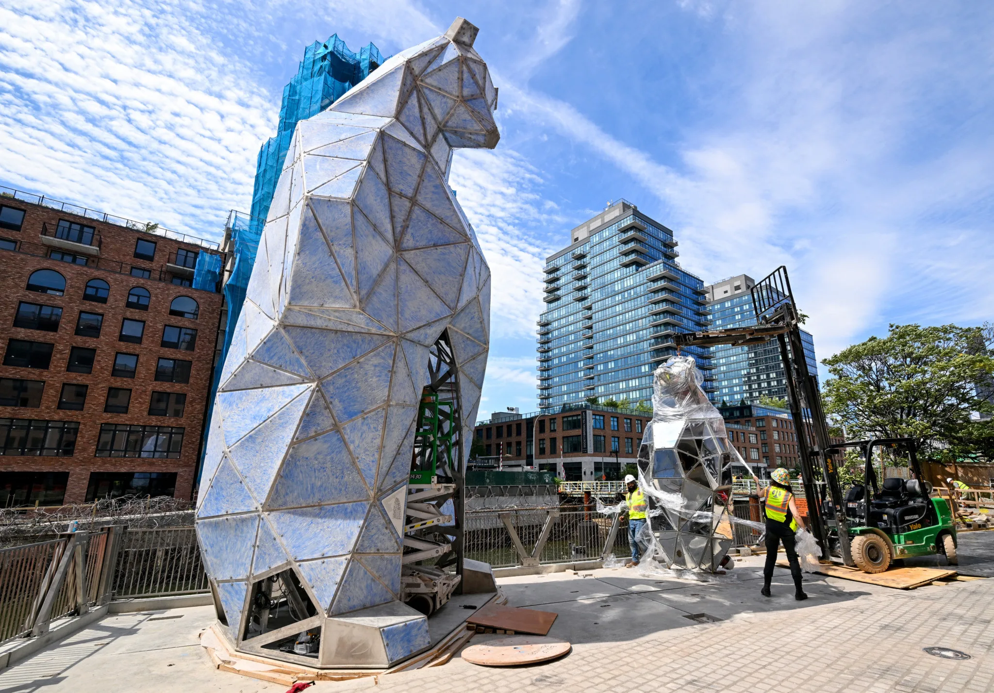 Construction workers installing large geometric bear sculptures in urban area with forklift and buildings.