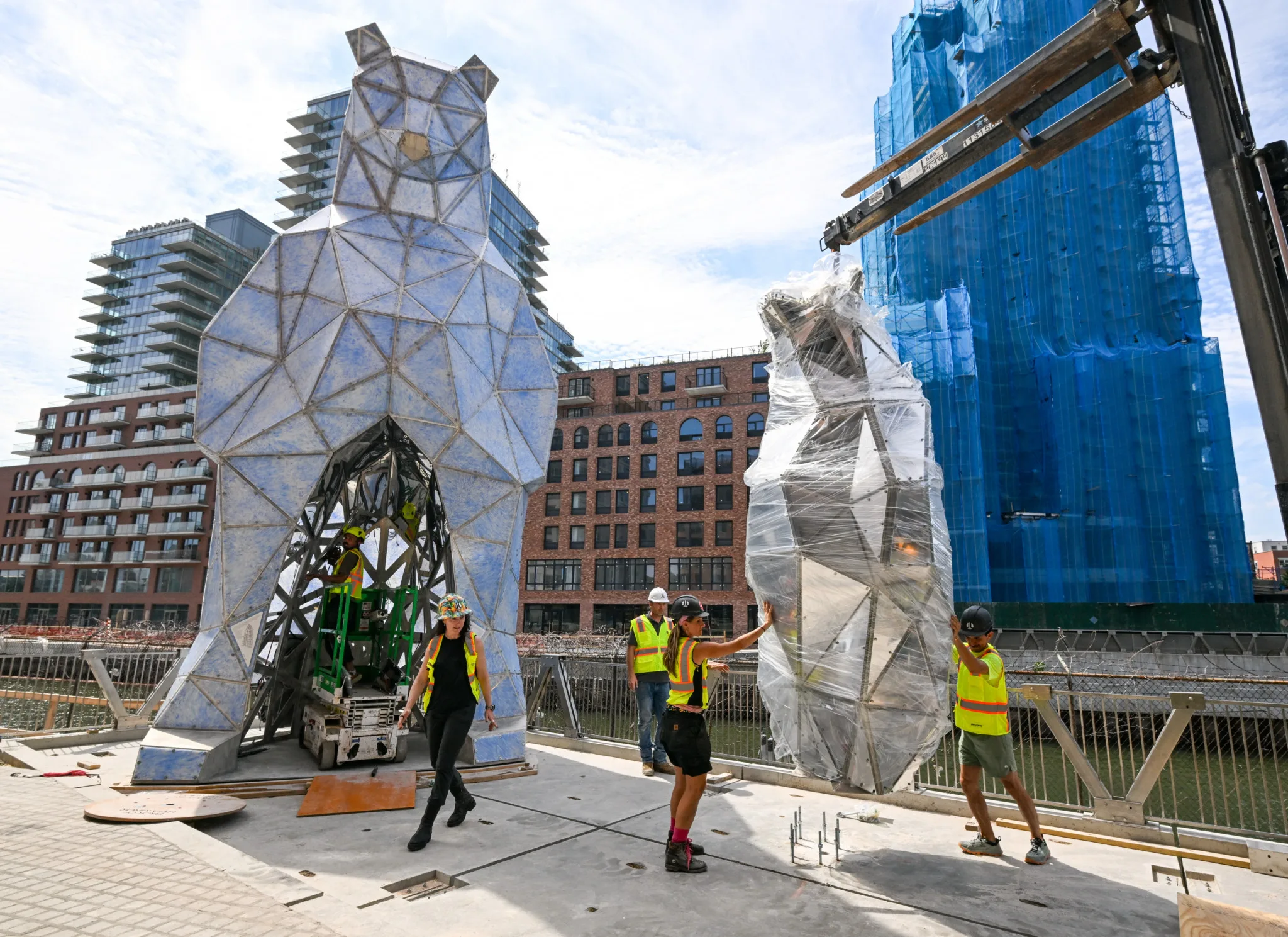 Workers in safety vests installing geometric sculpture next to another covered piece at waterfront site.