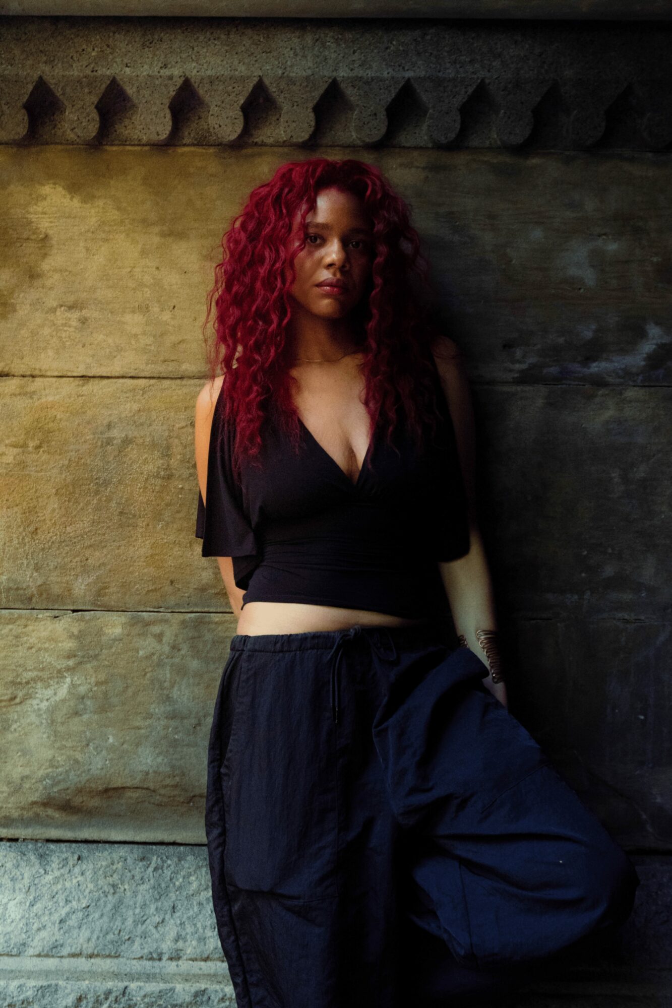 Woman with curly red hair standing against a textured wall, wearing a black top and loose pants.