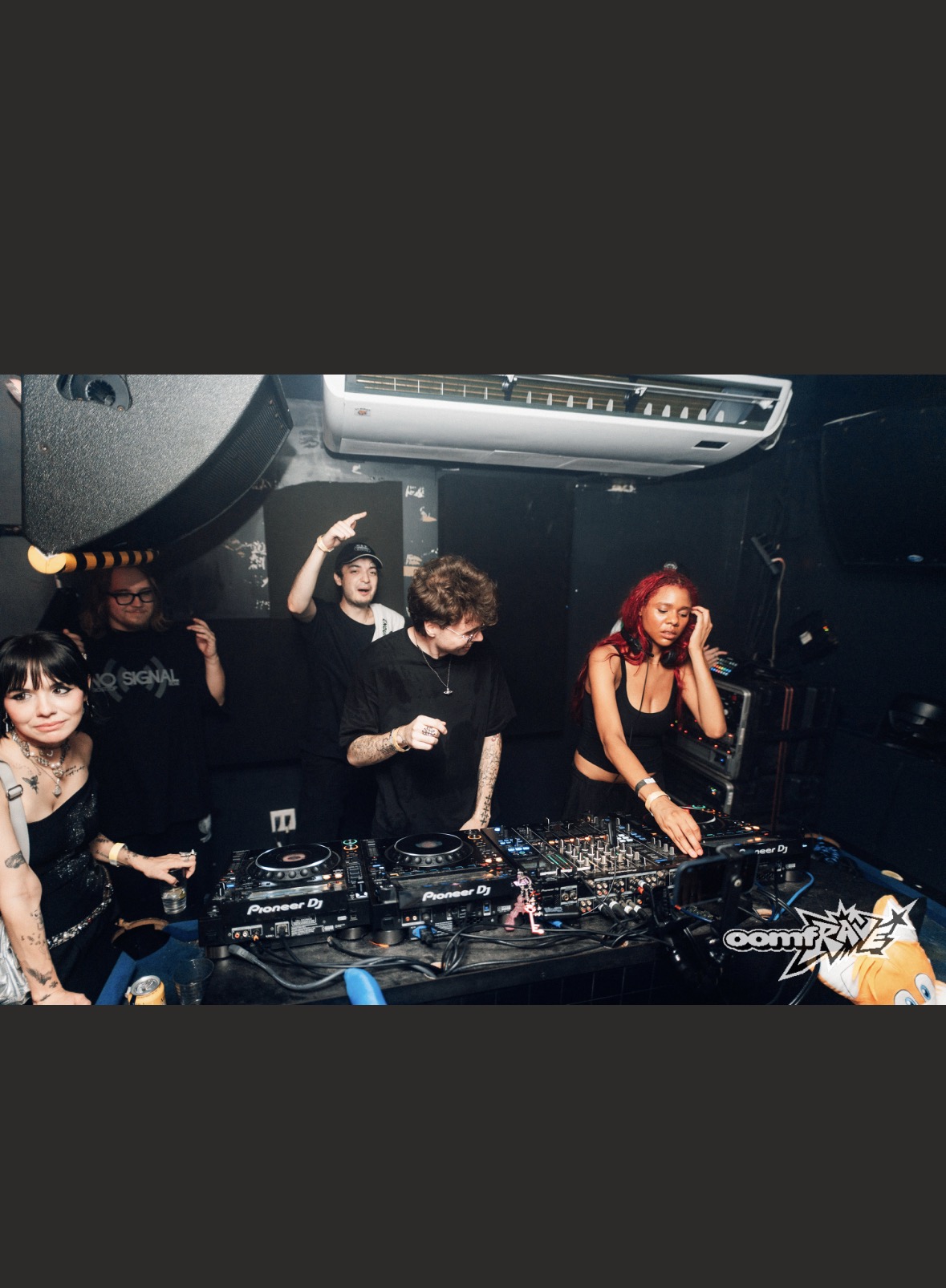 Group of people DJing at a club with equipment, some raising hands, one woman with red hair, dark background.
