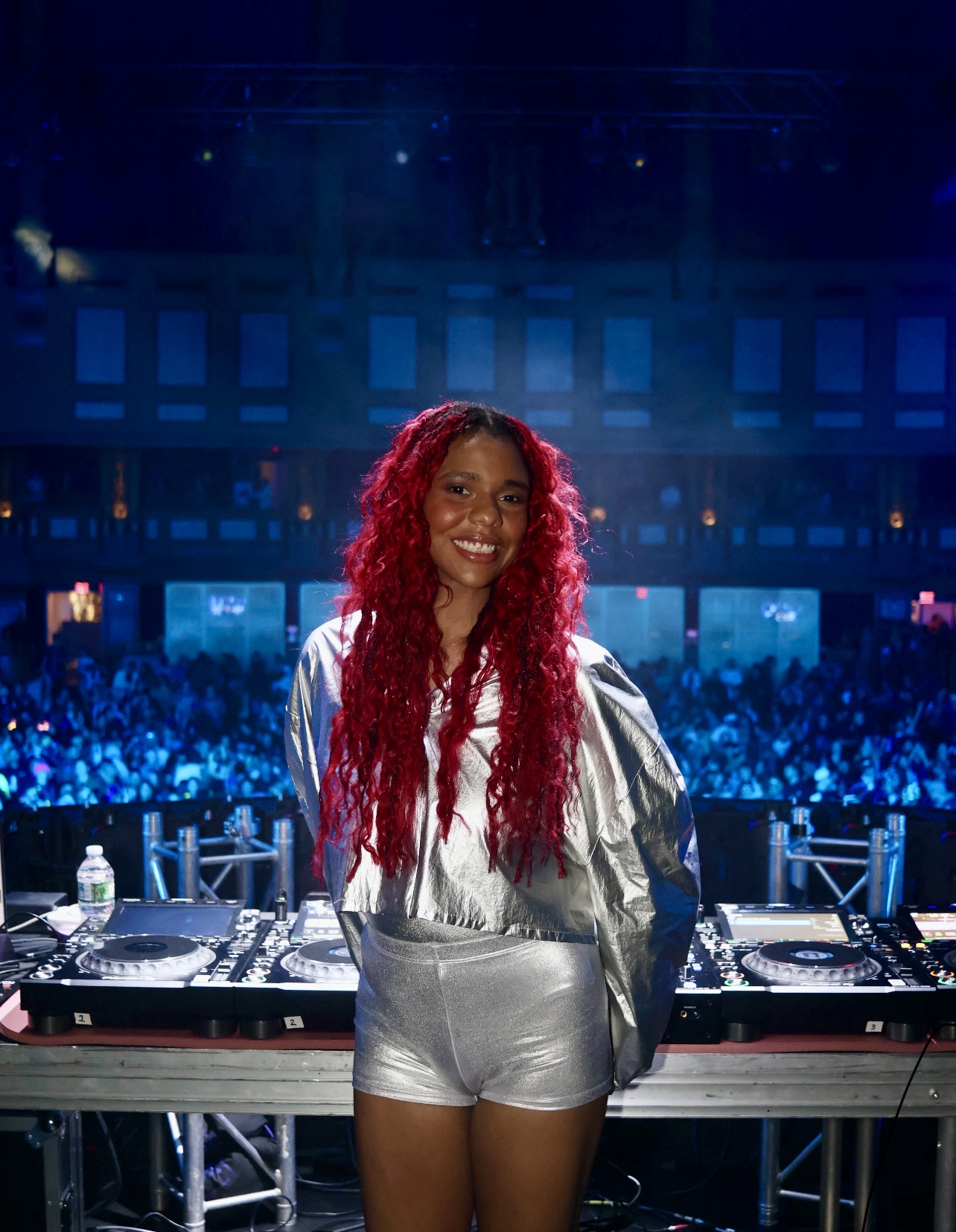 Smiling woman with long red hair standing behind DJ equipment in a large, illuminated concert venue.