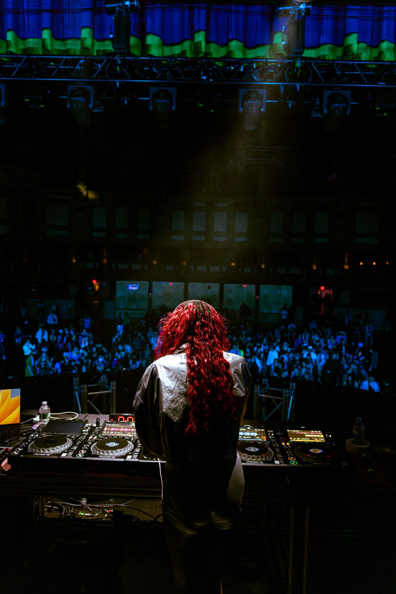 DJ with long red hair stands at turntables in a dark concert hall with a large audience and stage lighting.