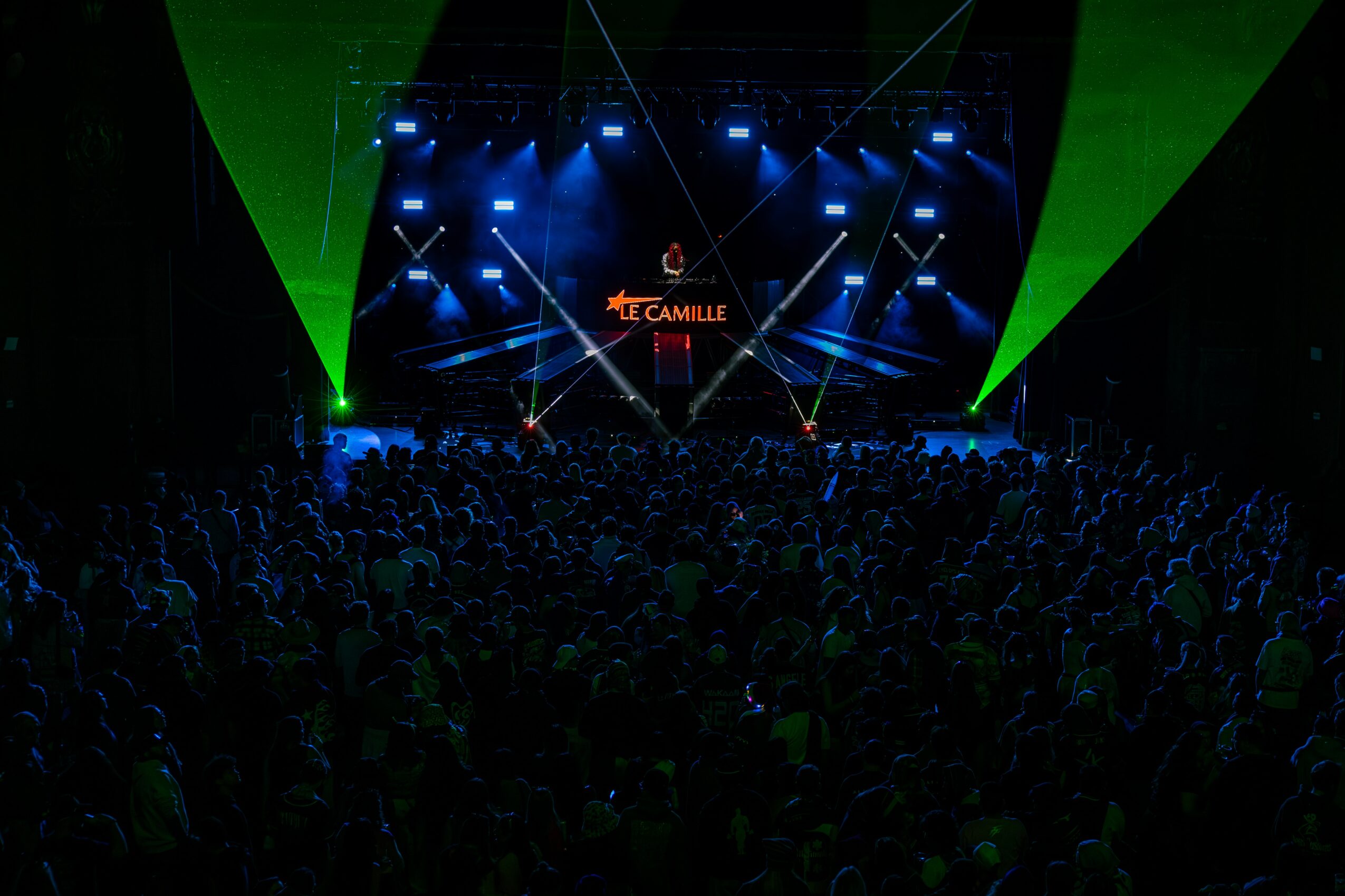 Concert stage with bright lights and audience in front, with green and blue lighting effects, and a sign reading 'Camille'.