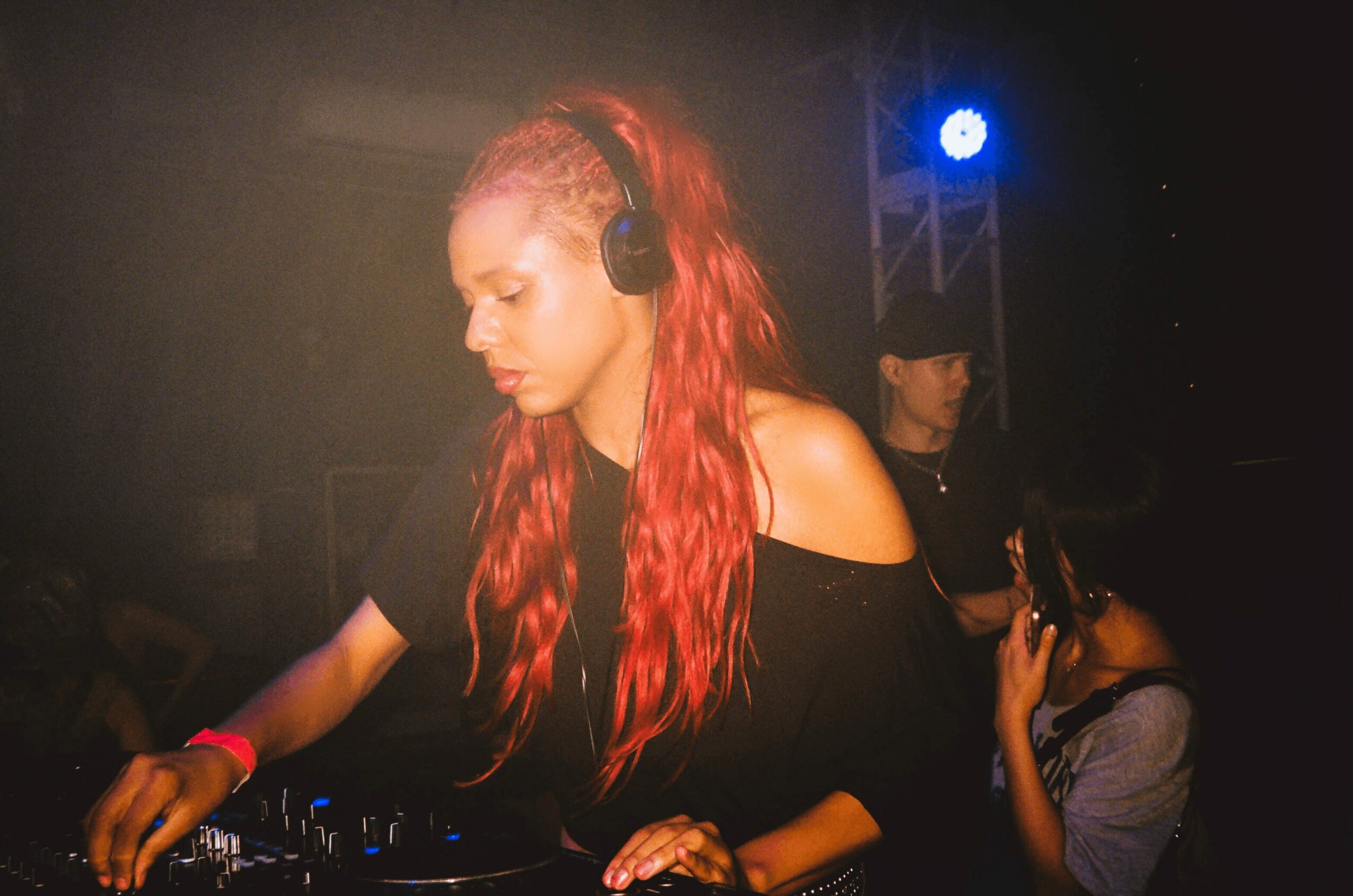 Female DJ with long red hair wearing headphones, focused on mixing music in a dark club setting.