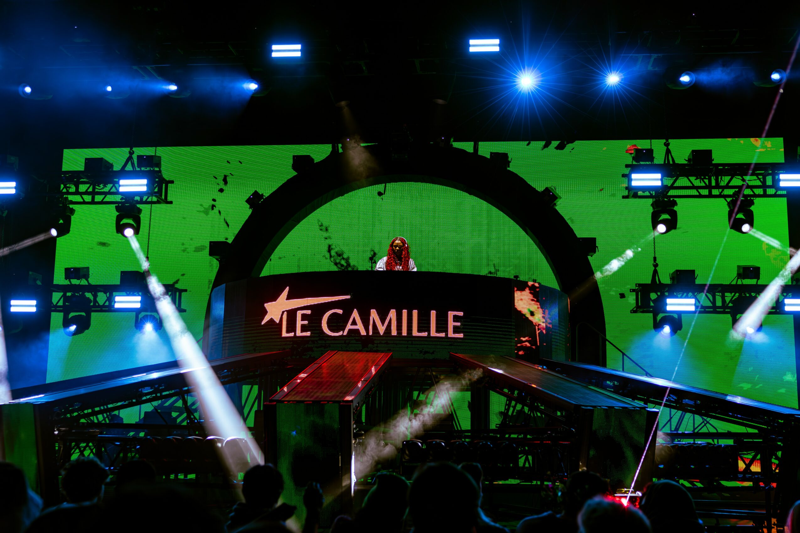 Stage with green background, bright lights, and a performer at a DJ booth, with the name 'Camille' displayed.