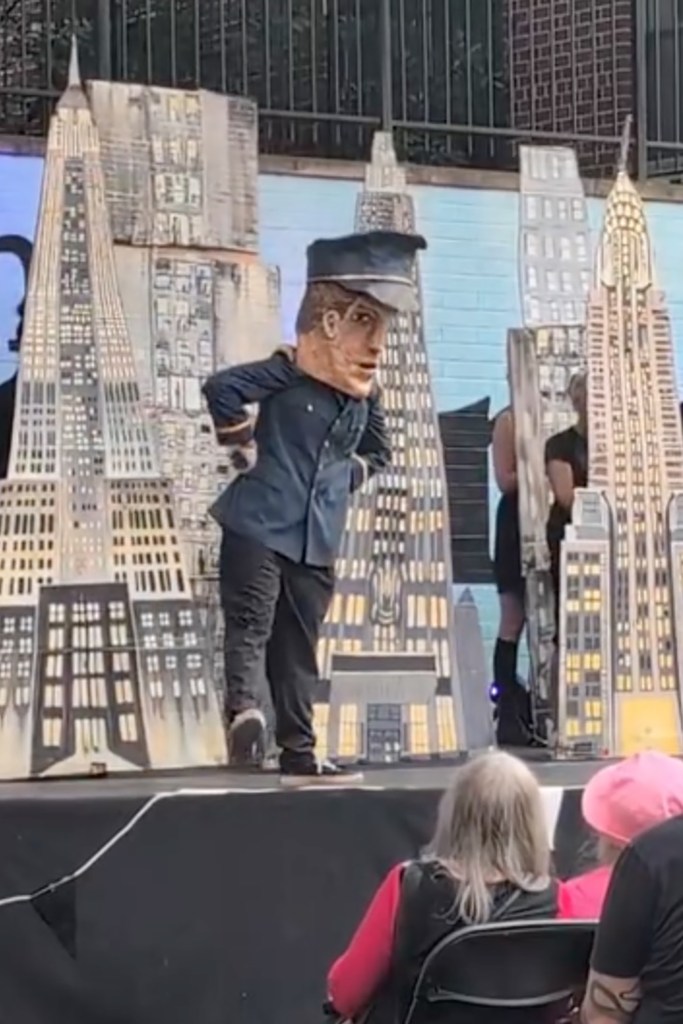 A Cardboard Pizza Puppet Circus performer in a large puppet costume, resembling a police officer, stands on stage surrounded by cardboard cutouts of city buildings during a performance.