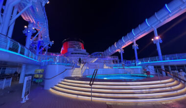 Aboard the Disney Wish, the cruise ship deck at night comes alive with a spiral staircase leading to an enchanting hot tub, vibrant lighting dancing around you, and thrilling water slides promising endless adventure.