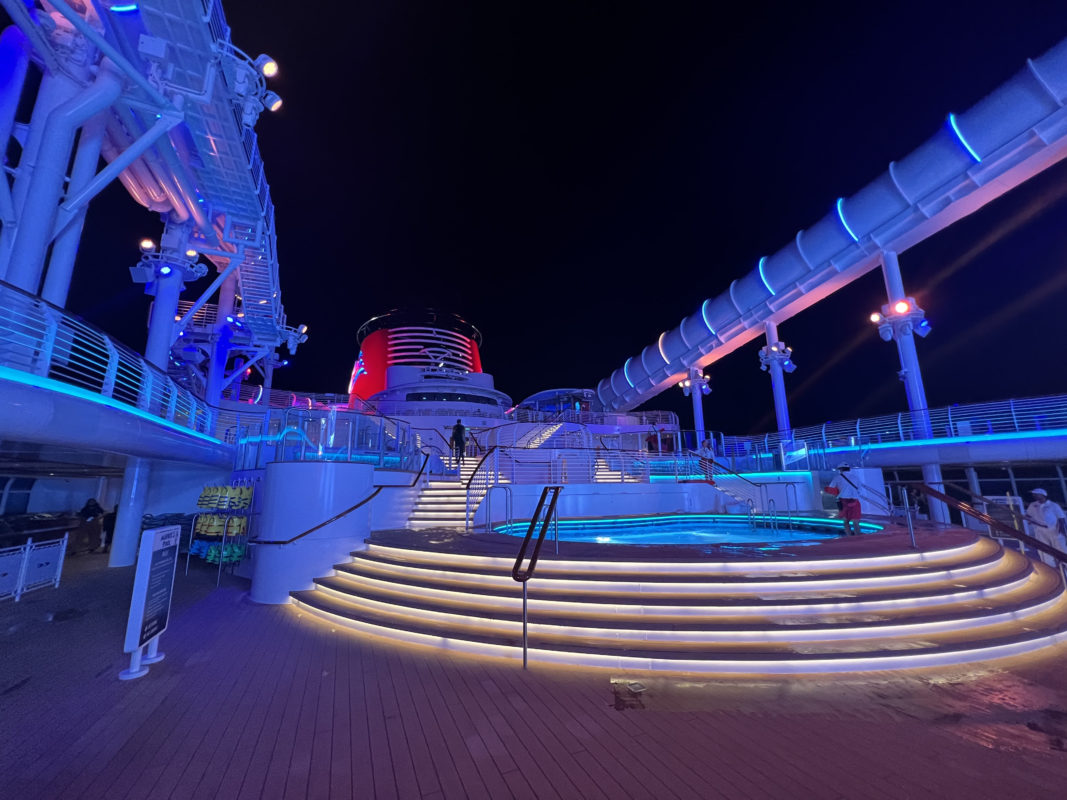 Aboard the Disney Wish, the cruise ship deck at night comes alive with a spiral staircase leading to an enchanting hot tub, vibrant lighting dancing around you, and thrilling water slides promising endless adventure.