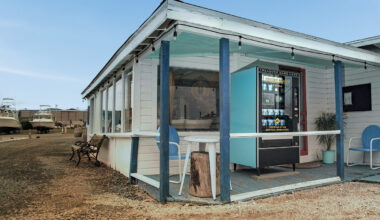 A vending machine stands on the porch of a small white building with blue trim. There are two blue chairs, a small table, and boats visible in the background. The area is rustic with gravel on the ground.