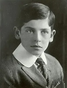 Black and white portrait of a young boy with slicked-back hair, wearing a patterned suit and a large collar.