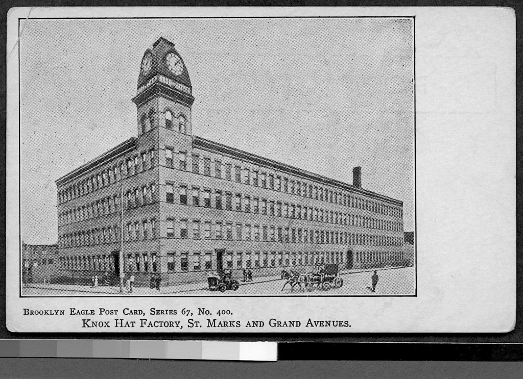 Black and white postcard image of the Knox Hat Factory, featuring a large brick building with a clock tower, situated at the intersection of St. Marks and Grand Avenues. The scene includes several people and a horse-drawn carriage in front.