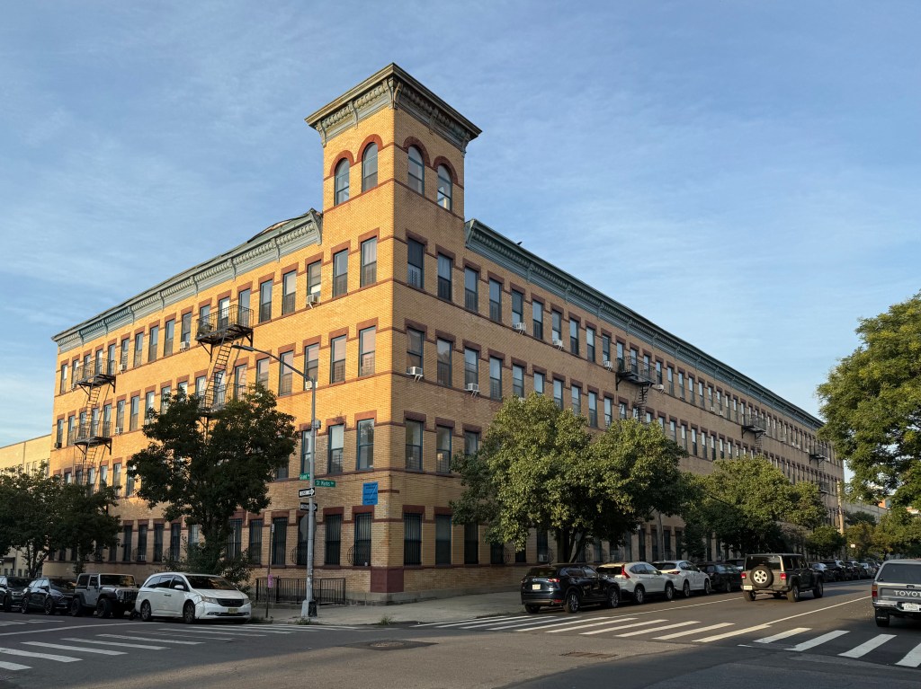 Historic brick building with multiple stories, featuring fire escapes and large windows, set on a city street with parked cars and trees.