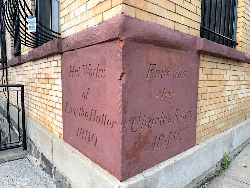 A brick building corner with an engraved stone featuring text that reads 'Hat Works of Knox the Hatter 1890' and 'Founded by Charles Knox 1840'.