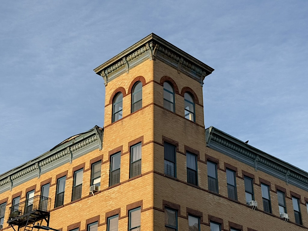 A close-up view of the top corner of a historic brick building with a tower featuring arched windows and decorative molding against a clear blue sky.