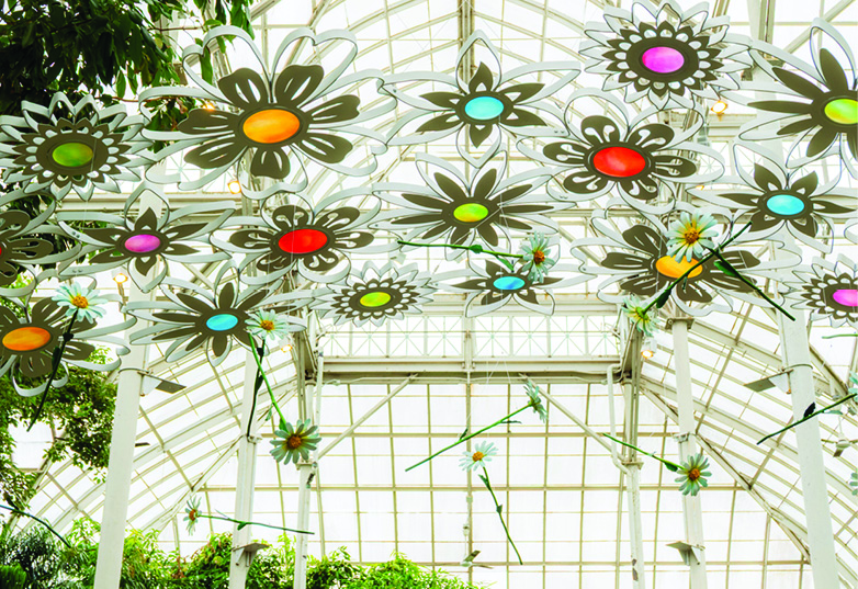 Colorful flower-shaped ornaments hanging from a glass greenhouse ceiling facade.