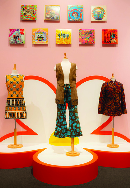 Three mannequins on a red-and-white curved platform display vintage outfits: left a sleeveless dress with geometric black and orange pattern, center a white top with brown vest and teal floral bell-bottoms, right a red-brown patterned jacket. A pink wall backdrop features two rows of colorful square artworks above the display.