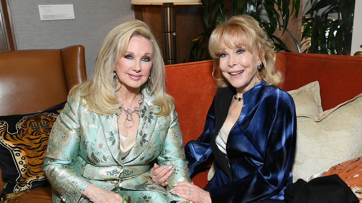 Morgan Fairchild wearing a silk light green suit sitting next to Barbara Eden wearing a blue stain blazer with a white blouse.