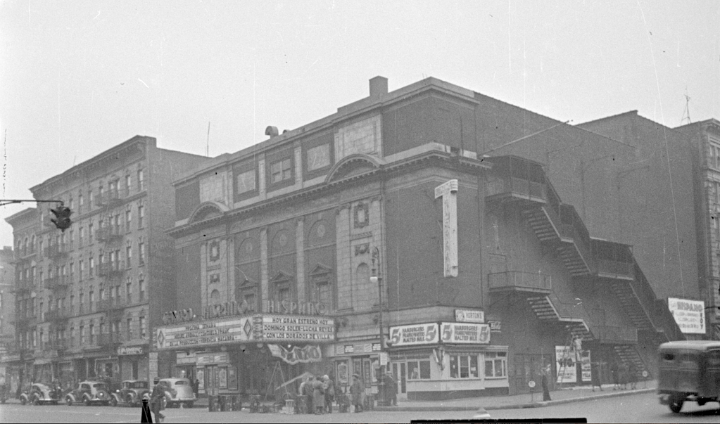 Historic theater building with marquee displaying movie titles, surrounded by urban architecture and traffic from the early 20th century.