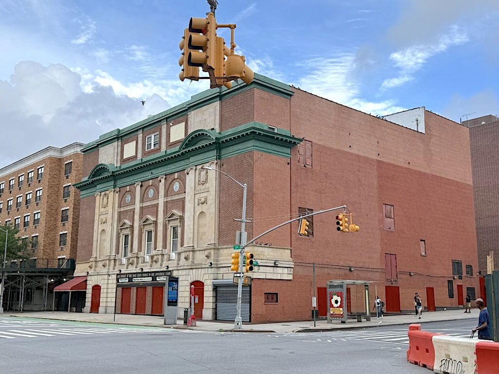 An urban scene showing a historic theater building with ornate architecture, featuring a green roof and decorative elements, surrounded by modern buildings, street lights, and pedestrians.