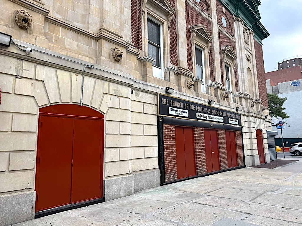 Exterior view of a building with red doors and decorative stonework, featuring a sign that reads 'The Church of the Lord Jesus Christ of the Lambs of God.'