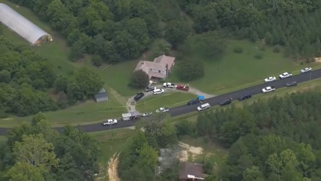 Aerial view of a house and surrounding rural area, with numerous emergency vehicles parked along the road and yellow crime scene tape visible.
