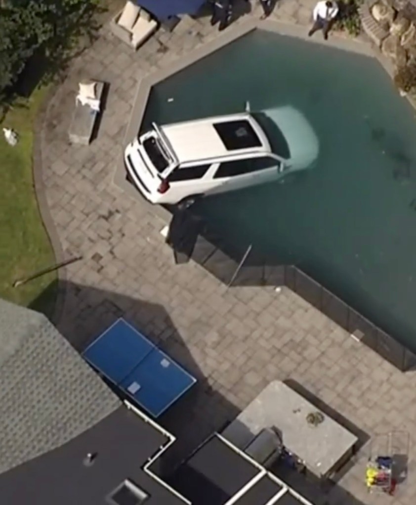 An aerial view of a white SUV partially submerged in a backyard swimming pool.