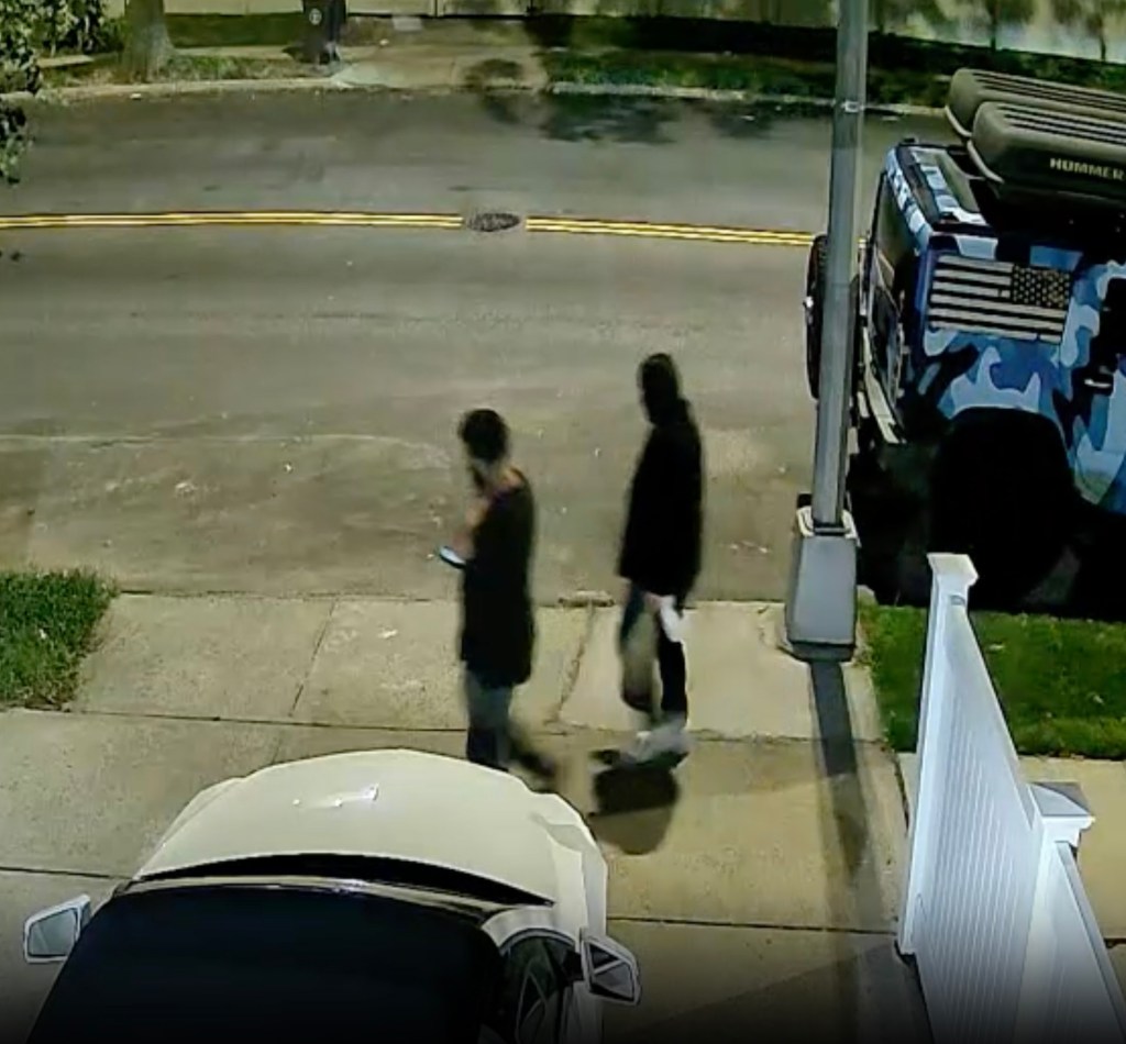 Security footage of two suspects walking on a sidewalk next to a vehicle.