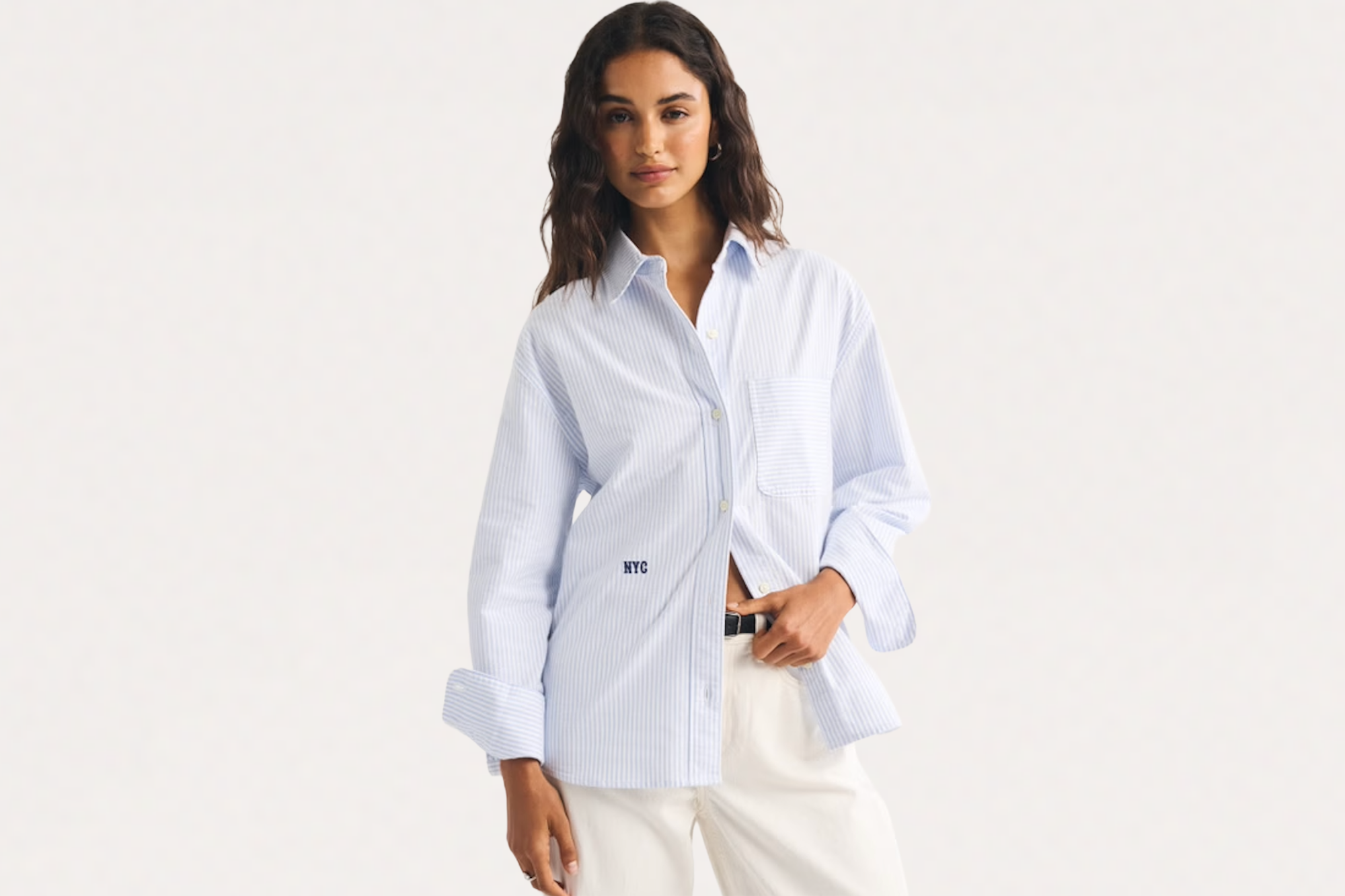 A woman wearing a light blue and white striped shirt with "NYC" embroidered on it, white pants, and a black belt.